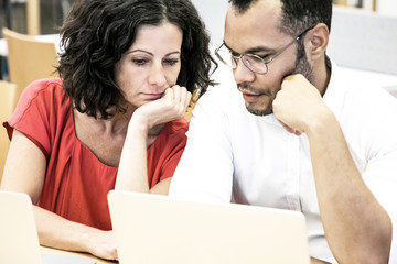 Tired adult students watching webinar in library. Man and woman in casual sitting at desk, looking at laptop monitor and talking. Online education concept