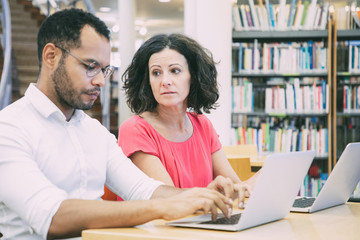 Wall Mural - Two adult college students working in library computer class. Man and woman in casual sitting at desk with laptops, typing and talking. Education and teamwork concept