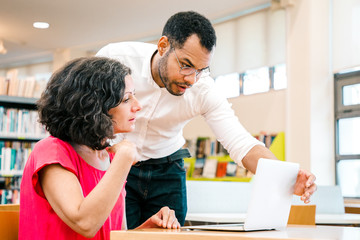 Wall Mural - instructor checking student work in library. man and woman in casual sitting and standing at desk, u