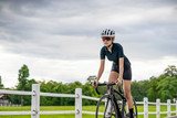 asian female bicyclist cycling pass a farm, standing up on the bicycle looking forward, riding a black bicycle wearing bike helmet and goggles, with grey clouds covering the sky and tall green trees.