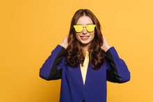 Happy, Stylish Girl In Yellow Glasses Touching Hair While Looking At Camera Isolated On Yellow