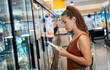 © Tijana - Woman grocery shopping stock photo. Young smiling woman shopping in local supermarket. She is shopping groceries. Standing by produce stand and reading nutrition label on dairy product.Choosing cheese