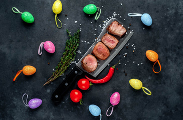 Wall Mural - Grilled beef steak over a meat knife with spices and decorative Easter eggs. View from above. on a stone background. Easter celebration concept