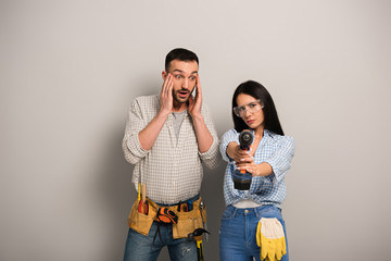 scared man looking at female manual worker with electric drill on grey