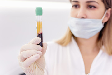 Wall Mural - Young attractive doctor with mask on face in scrub and gloves holds in hand test tube with separated plasma from the patient's blood. Blood plasma treatment, plasmolifting. Female medic professional