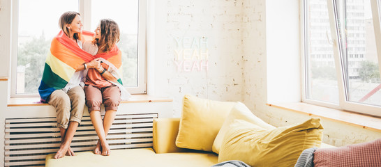 Beautiful female young lesbian couple in love huging near the window with the rainbow flag and holding each other's hands with the ring. Symbol of the LGBT community, equal rights.