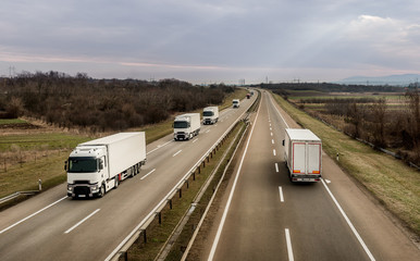 Convoy of transportation trucks passing on a highway on a bright blue day. Highway transportation with white lorry trucks