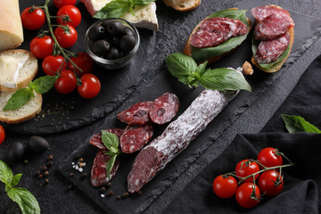 Wall Mural - The concept of Italian cuisine. Sausage on a black board with tomatoes, olives, basil and cheese. Bread with sausage, sandwich. Background image, copy space