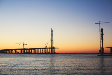 An unfinished part of highway road or bridge, on sunset or dusk, Industrial construction cranes and building silhouettes at sunrise, back light construction site silhouetted at sunset, over the water