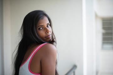 Wall Mural - Portrait of an attractive young brunette dark skinned Indian Bengali girl in western sportswear standing on a balcony  in white urban background. Indian lifestyle