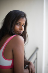 Wall Mural - Portrait of an attractive young brunette dark skinned Indian Bengali girl in western sportswear standing on a balcony  in white urban background. Indian lifestyle