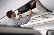 Young man putting luggage into overhead locker on airplane. Traveler placing carry on bag in overhead compartment