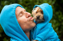 Portrait Of Dog Owner Kissing A Furry Friend In Matching Blue Hoodies Outdoors In Bright Green Park Background