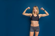 Portrait of beautiful athletic caucasian woman in sport bra and shorts showing biceps of her hand. Blue background. Copy space