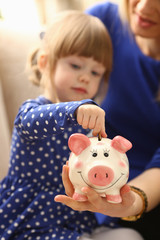 Child little girl arm putting pin money coins into happy pink faced piglet slot closeup. Making effective future needs savings collect dollar gift benefit present home leisure concept