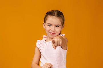 Cute smiling curly little girl in pink dress pointing camera on beige background. child pointing at you.