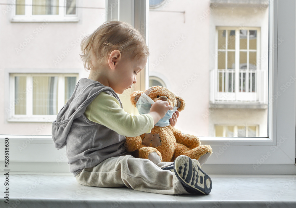 Child in home quarantine playing at the window with his sick teddy bear wearing a medical mask against viruses during coronavirus and flu outbreak. Children and illness COVID-2019 disease concept