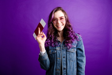 Bright girl with purple hair in pink glasses holding a passport with plane tickets in her hand