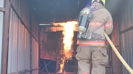 Wall Mural - Rear view of Fireman wear fire protection suit walking into the fire training room and Spray water Fire at fire station Slow Motion. Training fire drill Concept.