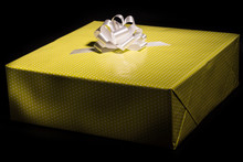 Large Gift Box Wrapped In Yellow Speckled Paper And A White Bow On A Black Background