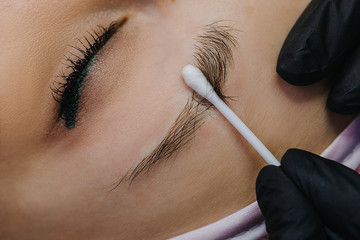 Wall Mural - Eye and eyebrow close up. A hand in a black glove holds a cotton swab and pushes the eyebrow hairs to see the first drawn hairs with microblading.