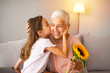 Cute girl giving a bunch of flowers to her grandmother sitting on the couch. Happy grandmother hugging small cute grandchild thanking for flowers presented. Darling granddaughter