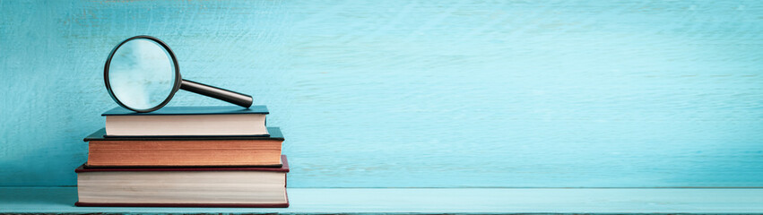 A magnifying glass on a stack of books on a blue wooden bookshelf with copy space web banner