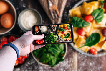 hands picking up a smartphone taking a picture of a fresh pasta on wooden table with ingredients. Space for text. Selective focus.