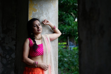 Young and beautiful Indian Bengali brunette woman is standing thoughtfully on the porch of a vintage house wearing Indian traditional ethnic vibrant skirt blouse. Indian lifestyle and fashion