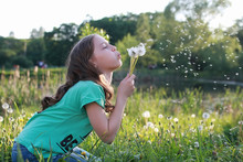 Teen Blowing Seeds From A Dandelion Flower In A Spring Park