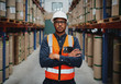 © StratfordProductions - Confident young manager with arms crossed standing wearing orange vest and white hardhat in manufacturing unit looking at camera