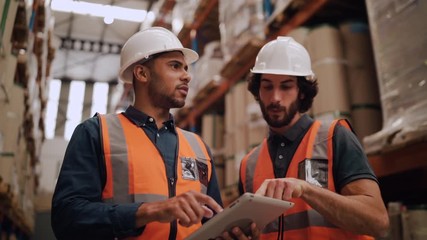 Diverse young male workers discussing and using digital tablet while supervising production at distribution warehouse