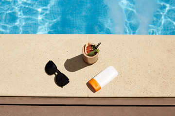 Summer holiday, vocation, relaxing, happyness, fashion concept. Woman's things at wooden pier at pool in spa, black sunglasses, sunscreen and cocktail near water in swimming pool at luxury resort.