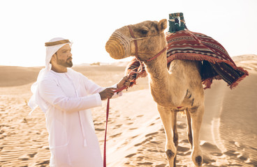 Wall Mural - Man wearing traditional clothes, taking a camel out on the desert sand, in Dubai