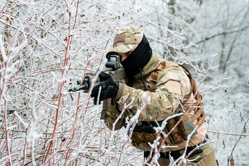 Wall Mural - Airsoft man in camouflage uniform and machinegun with optical sight. Soldier in the winter forest. Horizontal photo side view.