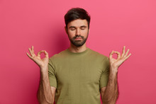 Relieved Bearded Young Man Relaxes During Meditation, Keeps Eyes Closed, Spreads Palms Sideways In Nirvana, Wears Casual T Shirt, Practices Yoga, Inhales Fresh Air, Isolated On Pink Studio Wall