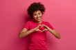 © WHstudio Leushin N - Pretty curly African American woman confesses in love, makes heart gesture, shows her true feelings, has happy expression, wears casual red t shirt, poses over pink background. Relationship concept