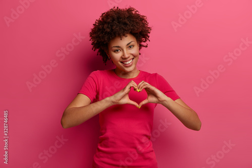 Fotografía Pretty curly African American woman confesses in love, makes heart gesture, shows her true feelings, has happy expression, wears casual red t shirt, poses over pink background