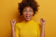 © wayhome.studio  - Yes, I did it! Euphoric dark skinned woman raises hands with clenched fists, looks happily, dressed in casual wear, stands against yellow background, feels like winner. People, emotions, lifestyle