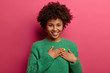 Pretty touched woman presses palms to heart, expresses positive feelings, feels touched to get help, makes gratitude gesture, wears warm green sweater, smiles sincerely, isolated on pink wall