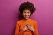© WHstudio Leushin N - Pleased girl in orange jumper presses palms to heart, makes thankful gesture, touched with cordial congratulations, smiles positively, isolated over purple background. Acknowledgement concept