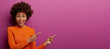 Leinwandbild Motiv Happy reaction on advert. Positive excited woman has Afro hairstyle, dressed in orange jumper, has widely opened eyes, shows blank space on purple background for your promotion or information