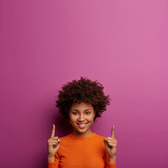 Wall Mural - Pretty cheerful African American girl invites you going upstairs, recommends product and points above, has happy expression, wears casual orange jumper, isolated on purple wall, gazes with joy