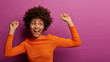 Positive carefree young woman with Afro hairstyle feels relaxed and happy, has fun at party, gazes aside, has beaming smile, isolated on purple background. People, hapiness, lifestyle concept