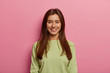 © wayhome.studio  - Portrait of attractive woman has healthy skin, has toothy smile, looks directly at camera, wears green jumper, has long straight hair, poses against pink pastel background. Face expressions concept