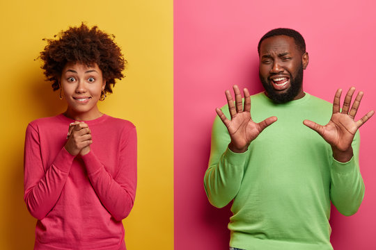 Emotive curly haired woman keeps hands together, gazes with anticipation, going to hear important results, displeased bearded man raises palms, discontent with something, poses over vibrant wall
