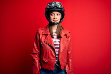 Wall Mural - Young beautiful brunette motocyclist woman wearing motorcycle helmet and red jacket smiling looking to the side and staring away thinking.
