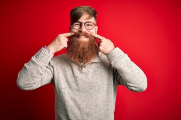 Canvas Print - Handsome Irish redhead man with beard wearing casual sweater and glasses over red background Smiling with open mouth, fingers pointing and forcing cheerful smile