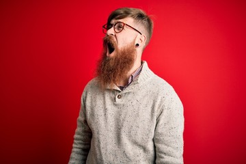 Poster - Handsome Irish redhead man with beard wearing casual sweater and glasses over red background angry and mad screaming frustrated and furious, shouting with anger. Rage and aggressive concept.