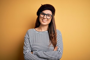 Young beautiful brunette woman wearing french beret and glasses over yellow background happy face smiling with crossed arms looking at the camera. Positive person.